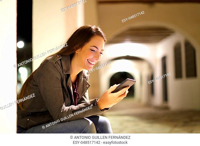 Happy woman using a smart phone sitting in the street in the night