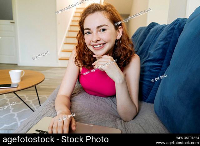Happy beautiful woman with laptop lying on sofa in living room at home