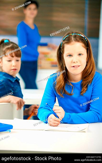Portrait of elementary age schoolgirl sitting at class looking at camera, with other student and teacher in background