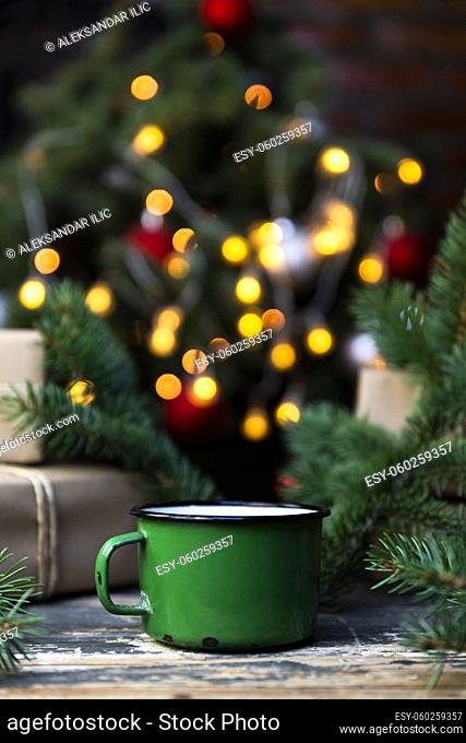 Coffee in a green tin mug on the rustic table with wrapped gifts, Christmas tree lights and spruce branches in the background