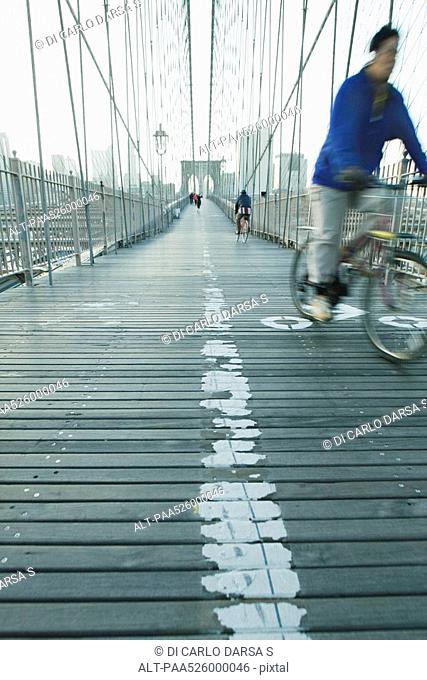 Man riding bike across pedestrian walkway of Brooklyn Bridge in New York City, Manhattan skyline in distance