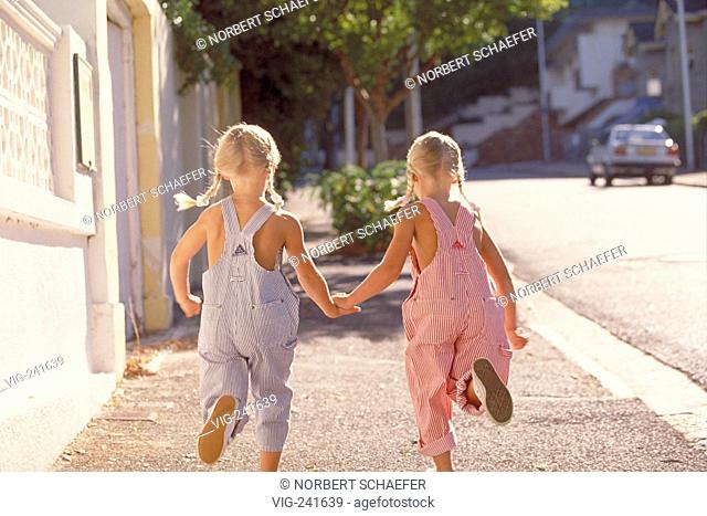 portrait, full-figure, two 5-year-old twin-girls with blond plaits wearing blue and red-white striped trousers with braces run hand-in-hand along the street  -...