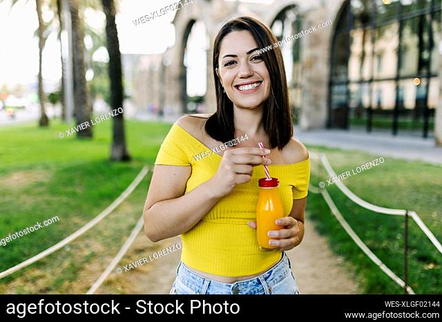 Smiling young woman holding orange juice bottle while standing in park