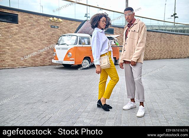 City style. Dark-skinned stylish girl in glasses sideways to camera and guy confidently looking at camera standing on city street