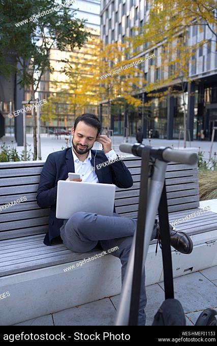 Businessman using in-ear headphones on retaining wall in city