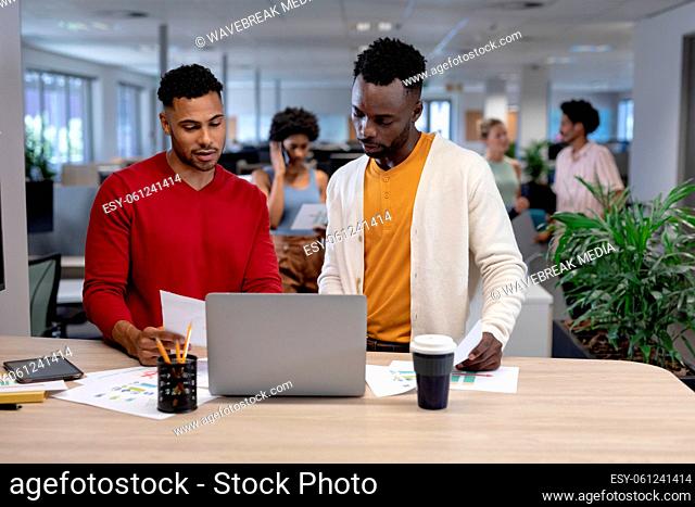 Biracial businessmen discussing over document with laptop at desk in modern workplace