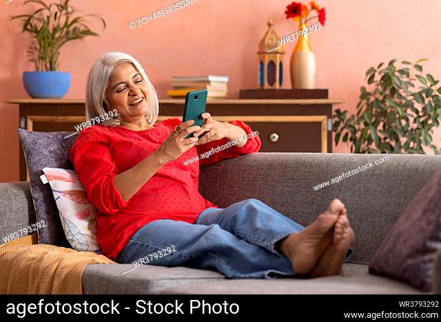 Old woman using mobile phone while relaxing on sofa in living room