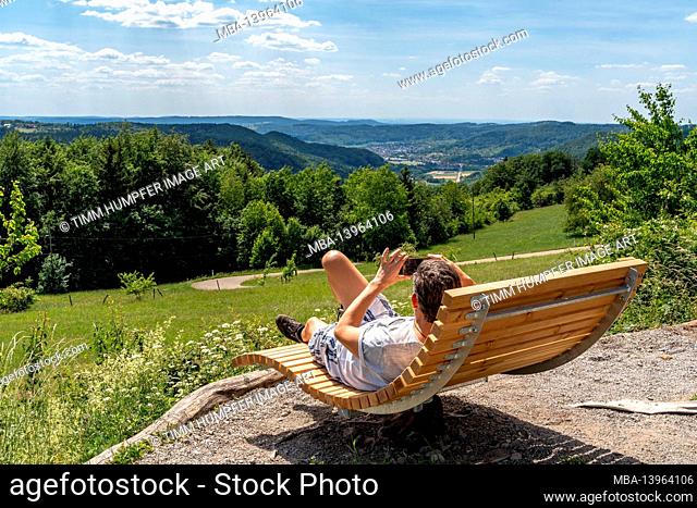 Europe, Germany, Baden-Wuerttemberg, Swabian-Franconian Forest Nature Park, Welzheim, hiker lies on a wooden lounger and takes a photo with the smartphone