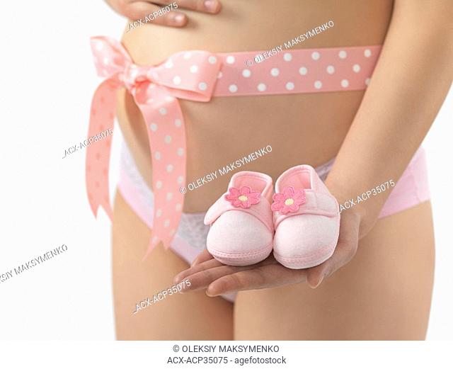 Pregnant woman with a pink bow on her belly holding baby shoes in her hand