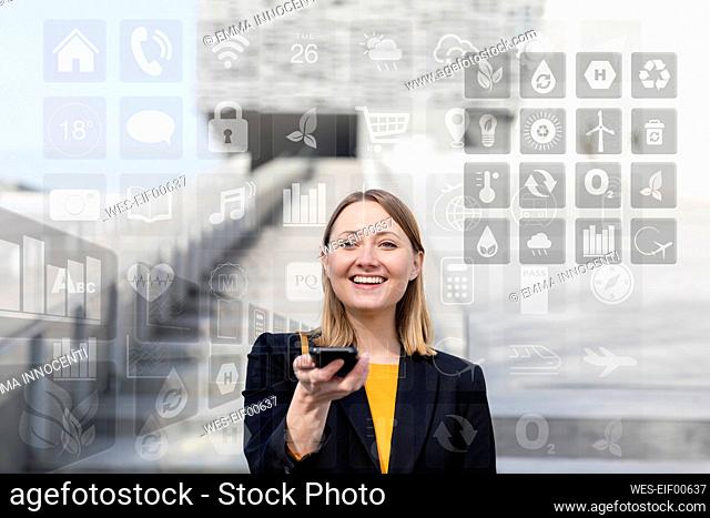 Smiling businesswoman holding mobile phone with different icons around outdoors