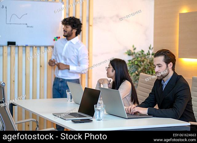 Company worker standing by the whiteboard in the meeting room while his coworkers sitting at the table