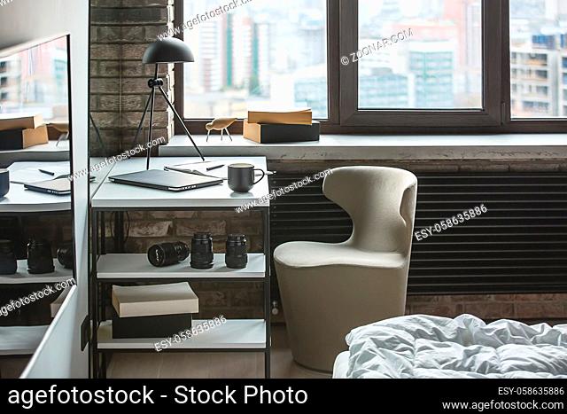 Room in a loft style with the brick wall and parquet on the floor. There is a bed with white blanket, armchair, white table with shelves