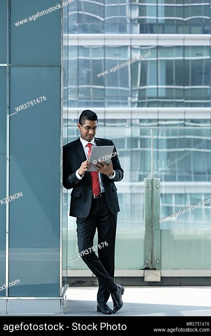 Indian businessman using a tablet PC while leaning on the wall of a modern office building indoors
