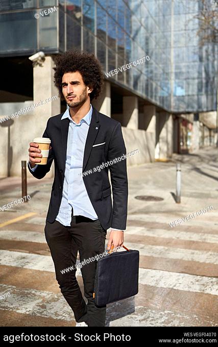 Businessman with laptop bag and coffee cup crossing road during sunny day
