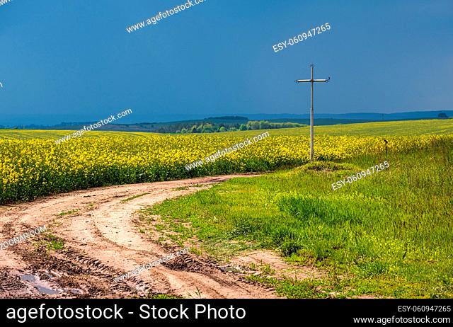 Cross near dirty road through spring rapeseed yellow blooming fields, cloudy sky. Natural seasonal, good weather, climate, eco, farming