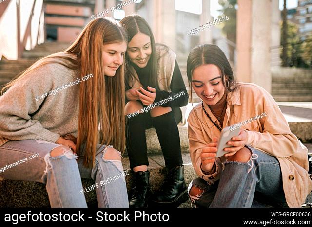 Smiling teenage girl showing mobile phone to friends while sitting on steps in city