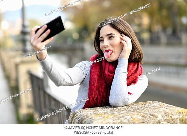 Portrait of young woman wearing red scarf sticking out tongue taking selfie with cell phone