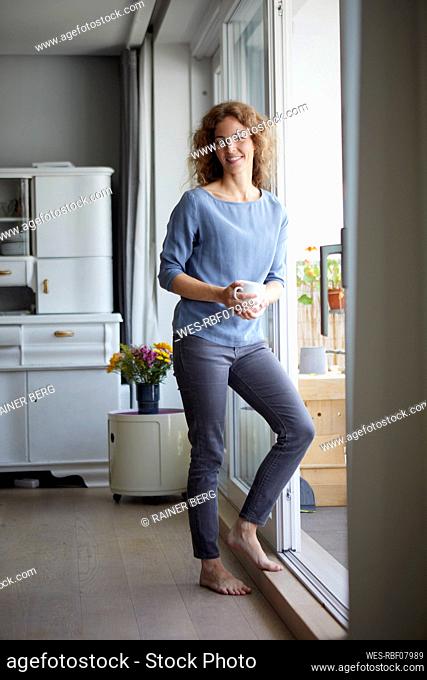 Smiling woman holding coffee cup while leaning on door by balcony at home