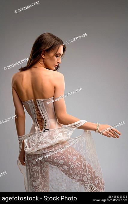 Confident seductive slim female model in translucent corset keeping hands on waist and undress peignoir
