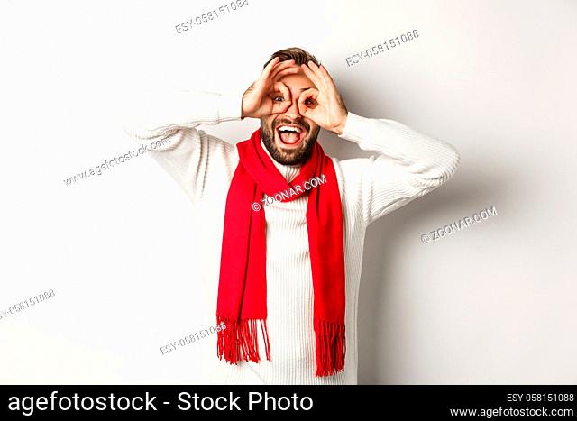 Happy guy celebrating christmas, showing funny faces and laughing, standing over white background in winter sweater and red scarf