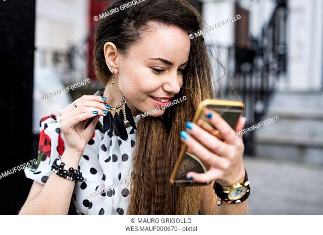 UK, London, young woman watching her earring with the aid of her smartphone