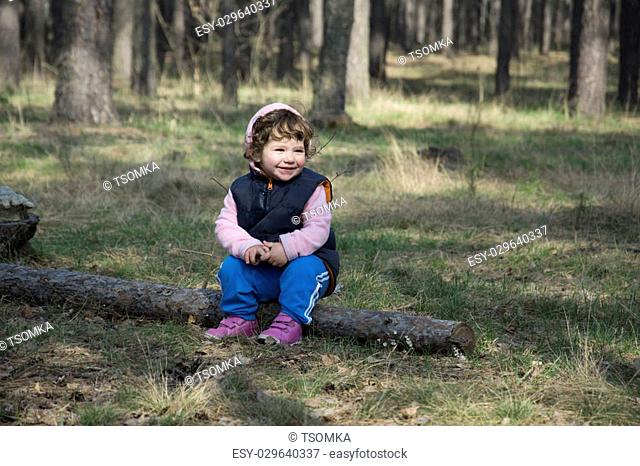 In the spring the little curly girl sitting on a log in a pine forest