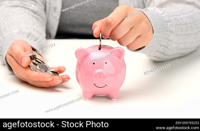 female hand throws a coin into a pink piggy bank on a white table. Concept of accumulating cash, saving, receiving subsidies