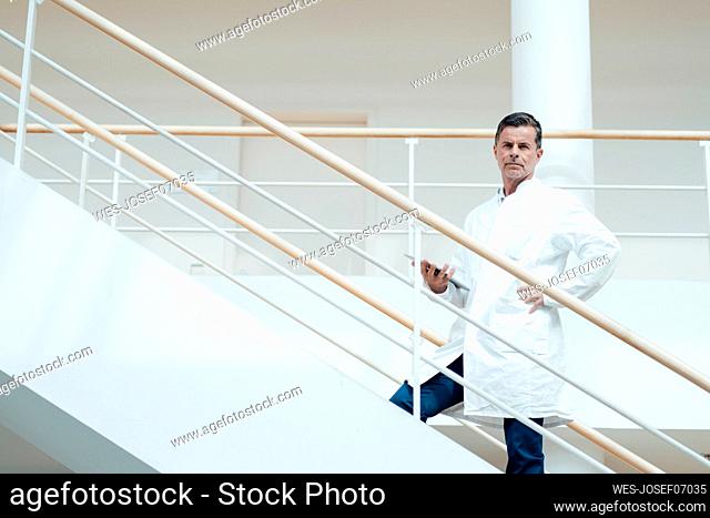 Serious scientist with hand on hip standing on staircase at medical clinic