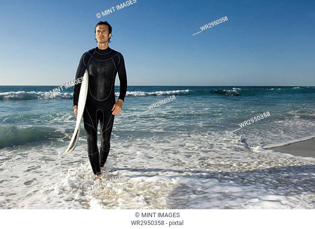 Surfer wearing wetsuit emerging from ocean, carrying surfboard