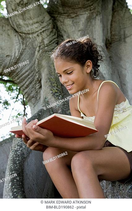 Hispanic girl reading book on tree trunk