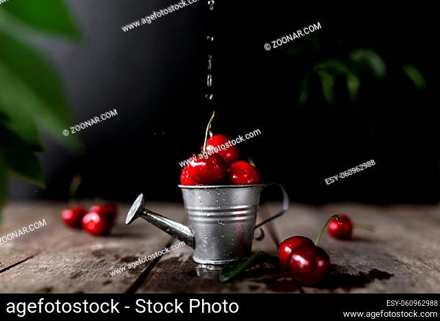 Wet sweet cherry berries in small watering can on wooden background table with flowing water stream and drops. Freshness, summer concept