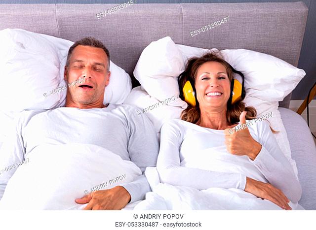 Happy Mature Woman Covering Her Ears With Headphone Showing Thumb Up Sign While Man Snoring