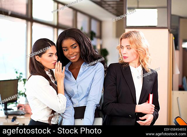 Three ladies standing in office. Two gossiping and blonde girl looking at them