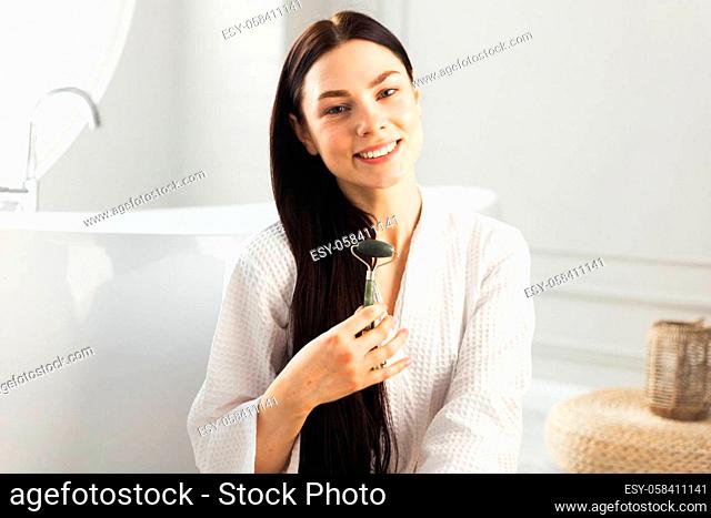 Beautiful girl holding drainage massage face roller sitting in luxury bathroom