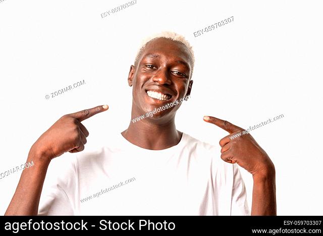 Close-up of pleased handsome african-american blond guy pointing at his white perfect smile, recommend dentist, being happy, standing over white background