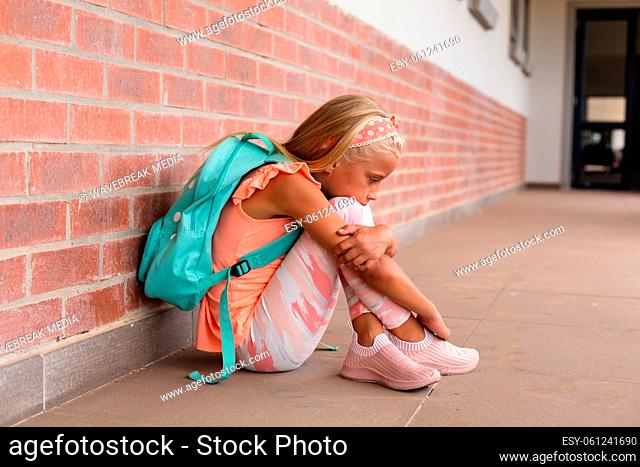Full length of sad caucasian elementary schoolgirl hugging knees while sitting on floor in school