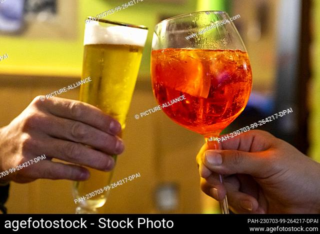 PRODUCTION - 01 July 2023, Berlin: In a bar, two visitors toast with a glass of non-alcoholic beer and a glass of Aperol Spritz