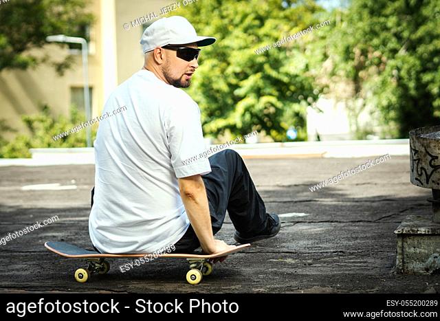 man in a white T-shirt is sitting on a skateboard and looks around on a summer day