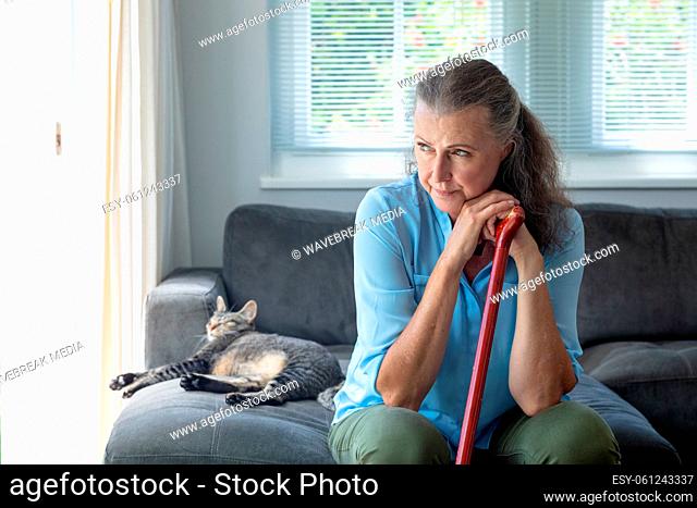 Thoughtful senior woman leaning on walking cane sitting by cat on sofa at home
