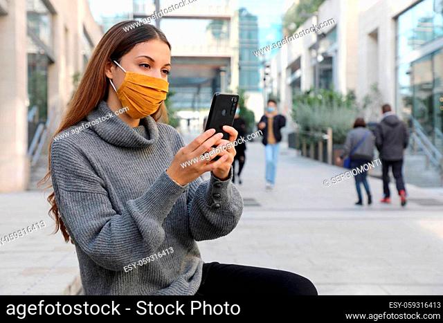 Attractive young business woman with face mask sitting on bench of pedestrian city street using a smartphone