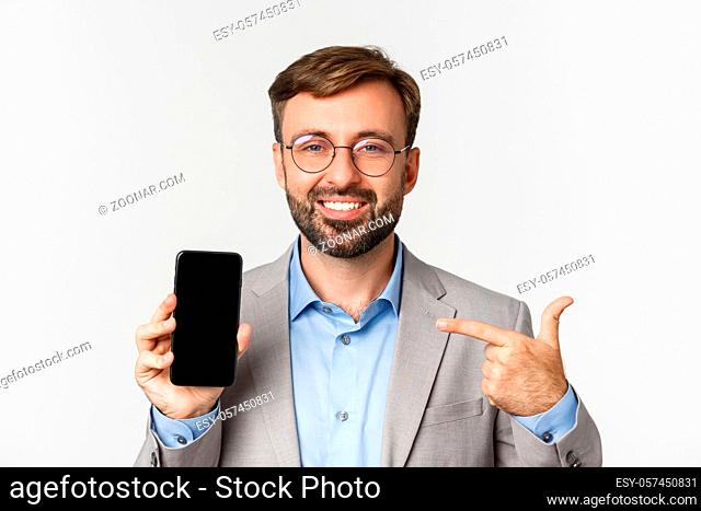 Close-up of confident businessman in glasses and gray suit, pointing finger at mobile phone screen and smiling proud, standing over white background