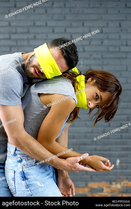 Blindfolded man leaning on woman while standing by gray wall
