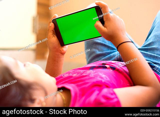 Little Indian Kid with phone in her hands sleeping on bed, mock up with green screen, focus on phone