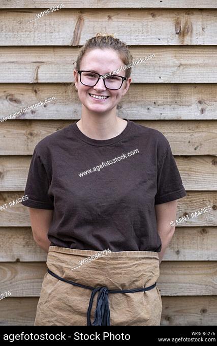 Portrait of female barista with blond hair and glasses, wearing brown apron, leaning against wooden wall, smiling at camera
