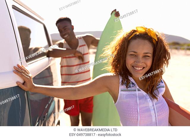 Front view of happy young Mixed-race couple with surfboard leaning on camper van at beach
