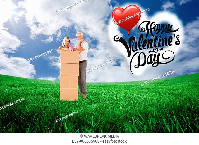 Composite image of happy couple leaning on pile of moving boxes with piggy bank