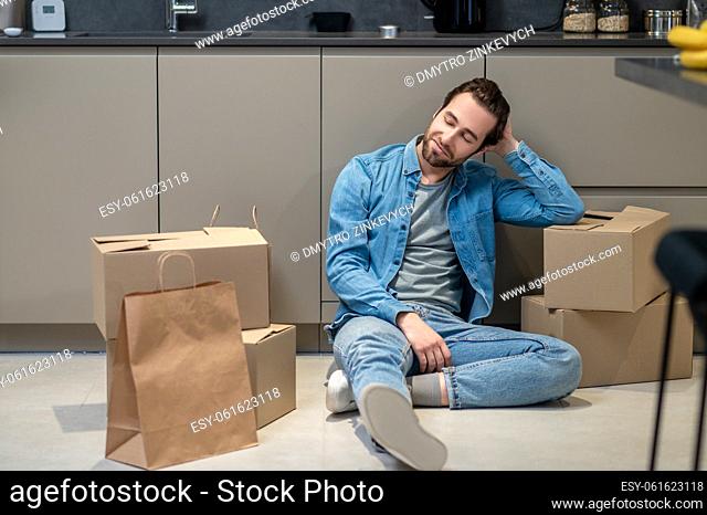 Moving, rest. Smiling young bearded man looking thoughtfully at bags and boxes sitting on floor in kitchen of new apartment