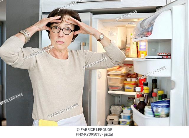 Portrait of an elderly woman looking shocked in front of a refrigerator