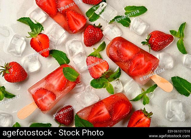 Close up fruit ice cream popsicle with fresh strawberry, green mint leaves and ice cubes on white table surface, elevated top view, directly above