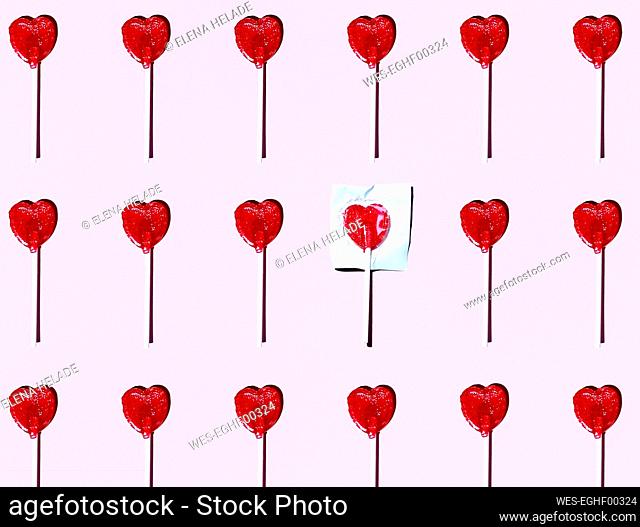 Pattern of heart shaped lollipops flat laid against pink background with single one still in wrapping paper